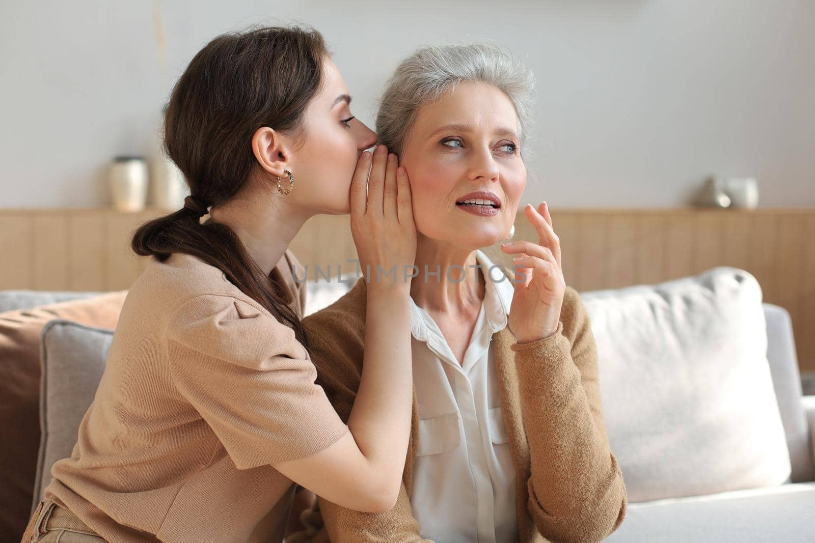Happy adult daughter whispering secret to her middle mother at home, gossiping, sharing secret. Beautiful woman hearing good news from mature mum, trusted relations.