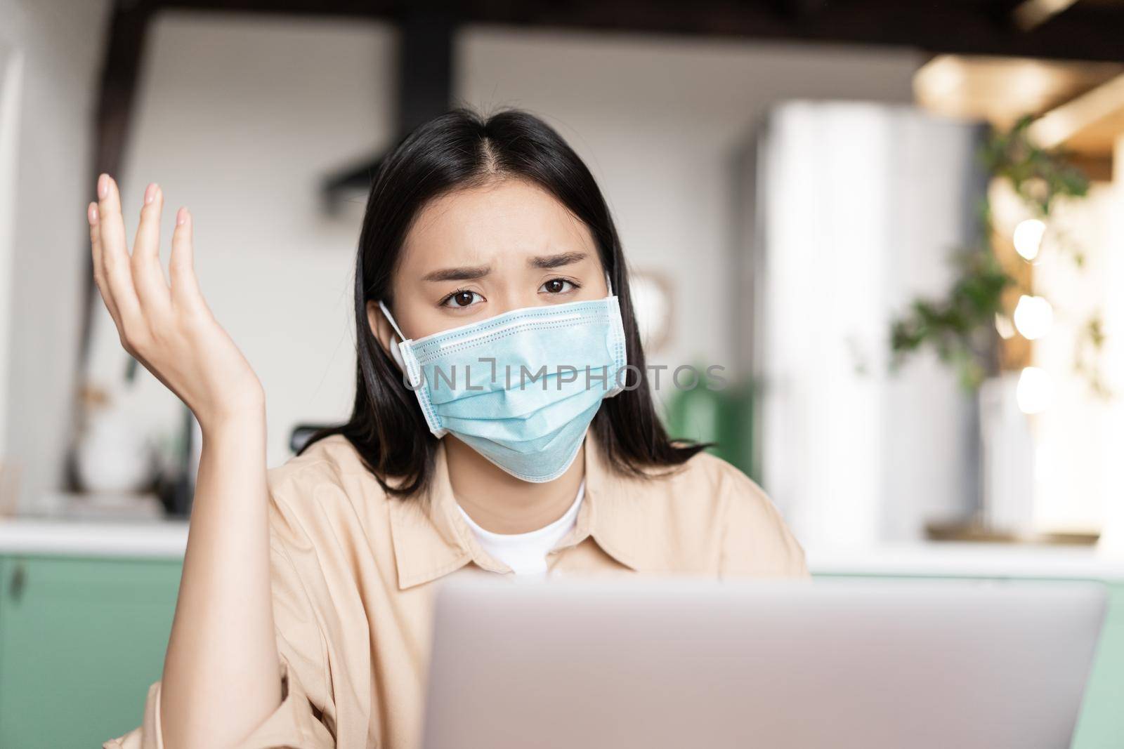 Troubled asian girl feeling sick, wearing medical mask, working on laptop by Benzoix