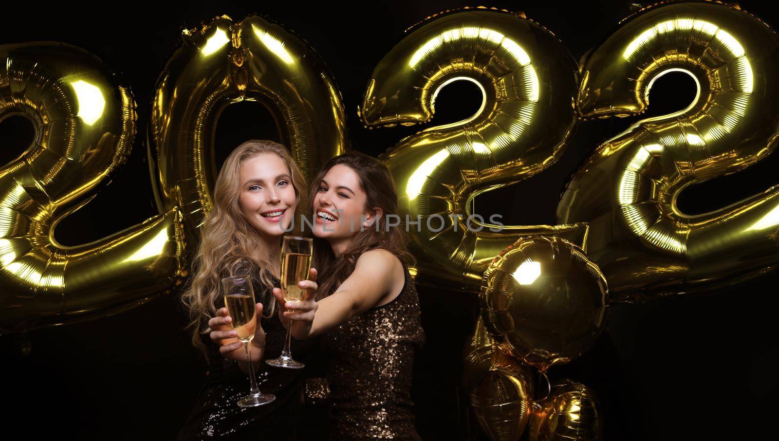 Two young ladies drinking champagne. Image of girls with balloons isolated on black background, having fun at Christmas or New Year's 2022 Eve Party