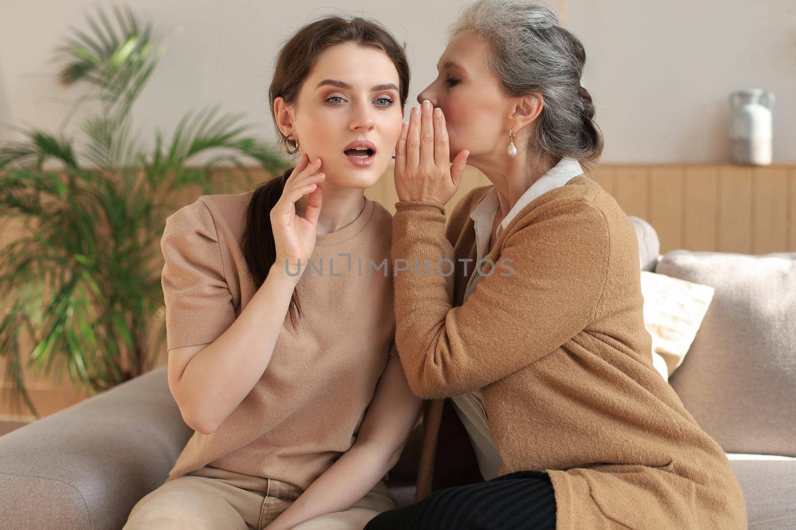 Happy middle mother whispering secret to her smiling daughter at home, gossiping, sharing secret. Beautiful woman hearing good news from mature mum, trusted relations
