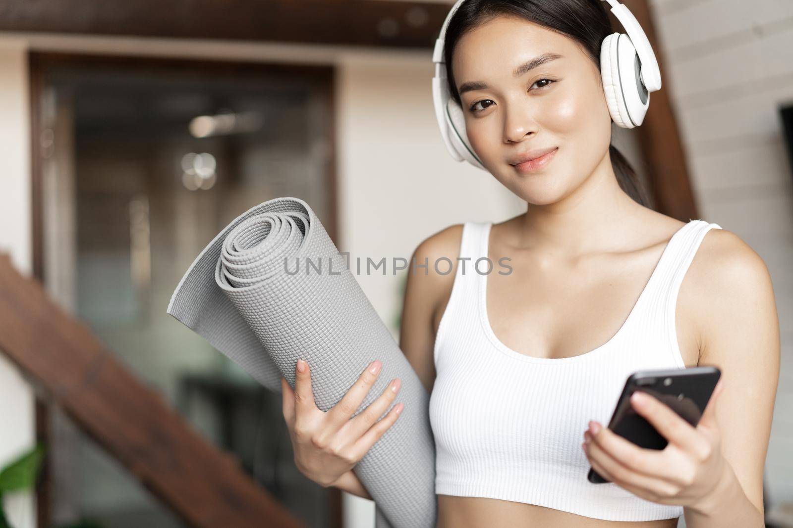 Smiling asian girl in headphones, listening music at home and workout with floor mat, holding mobile phone, fitness exercises in living room by Benzoix