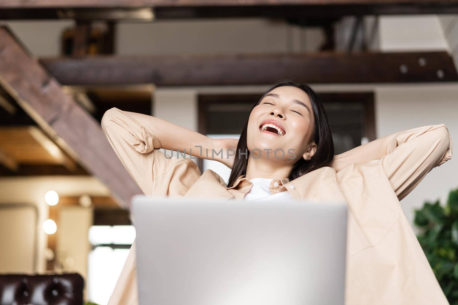 Smiling asian girl relaxing with laptop at home, lay back with hands behind head and pleased face expression.