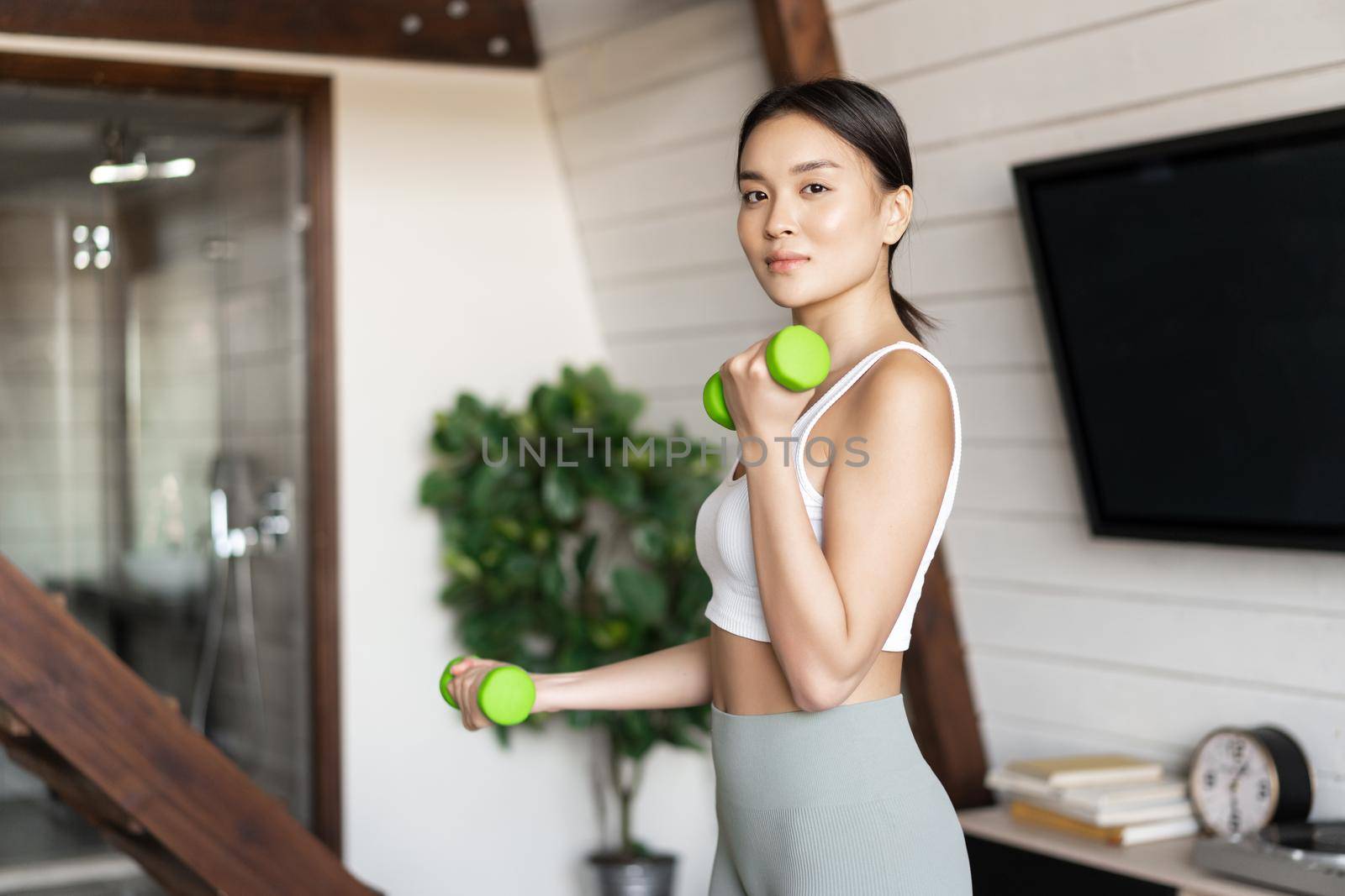 Active and healthy asian girl with fit body doing fitness exercises at home, lifting dumbbells and smiling, workout in living room by Benzoix