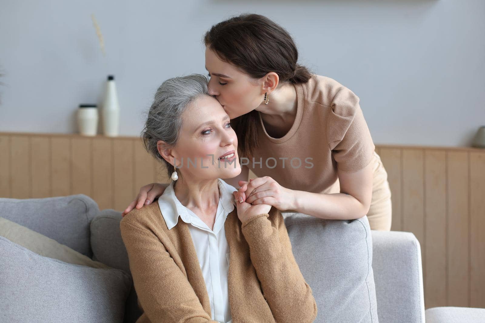 Happy daughter hugging older mother, standing behind sofa in living room, enjoying tender moment at home
