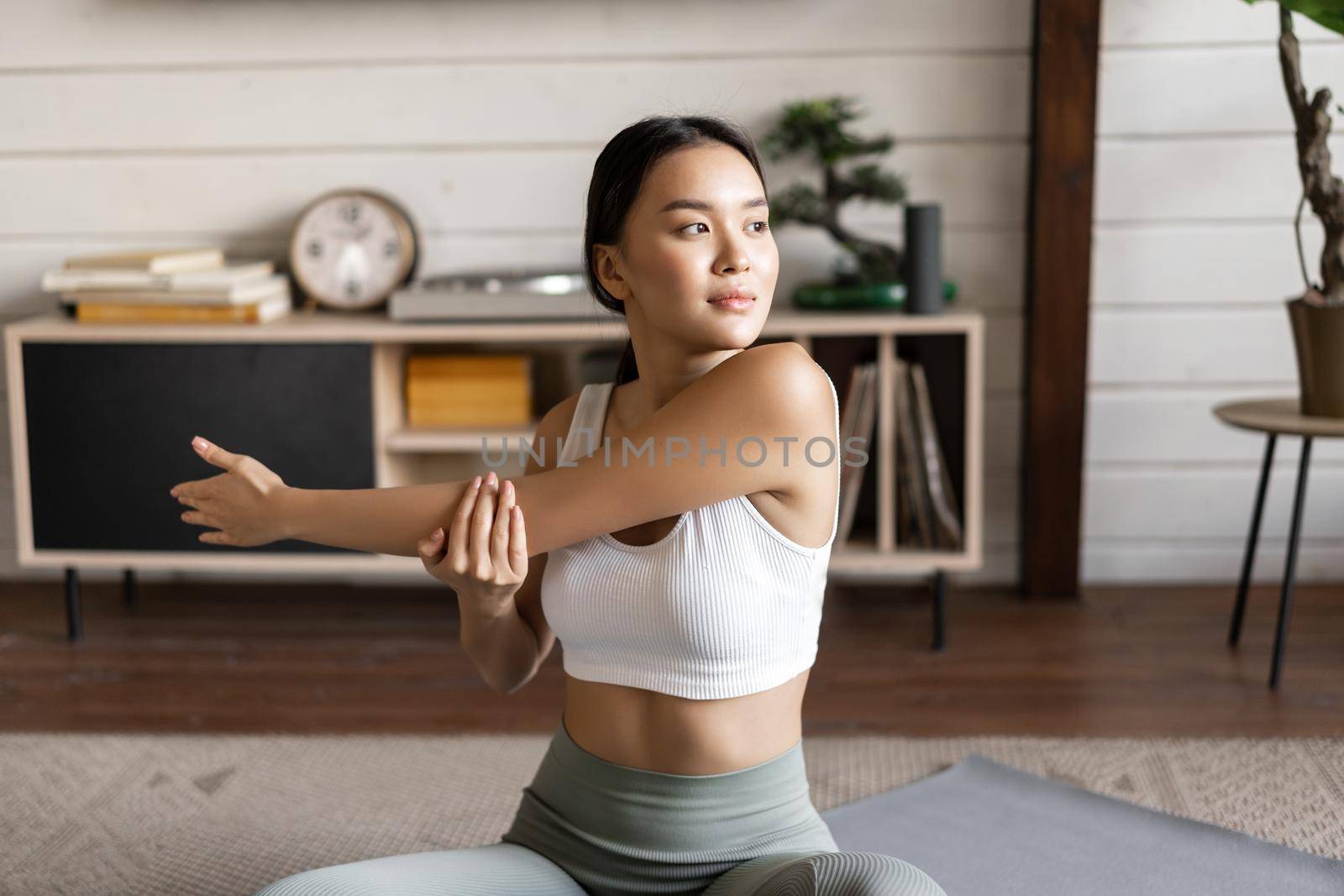 Asian fitness girl doing workout at home, sitting on floor mat in activewear, warm up and stretching arms, doing yoga in living room by Benzoix