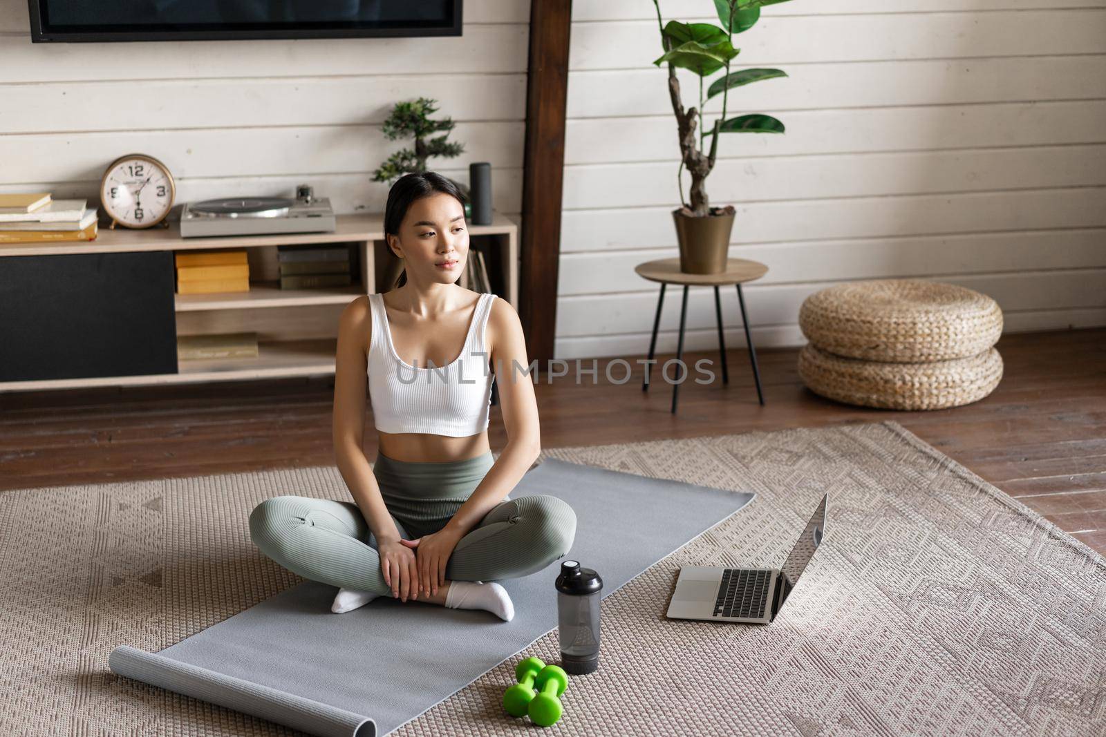 Asian fitness girl doing workout at home with laptop guide, sport application on computer, sitting on floor mat in living room, wearing activewear.