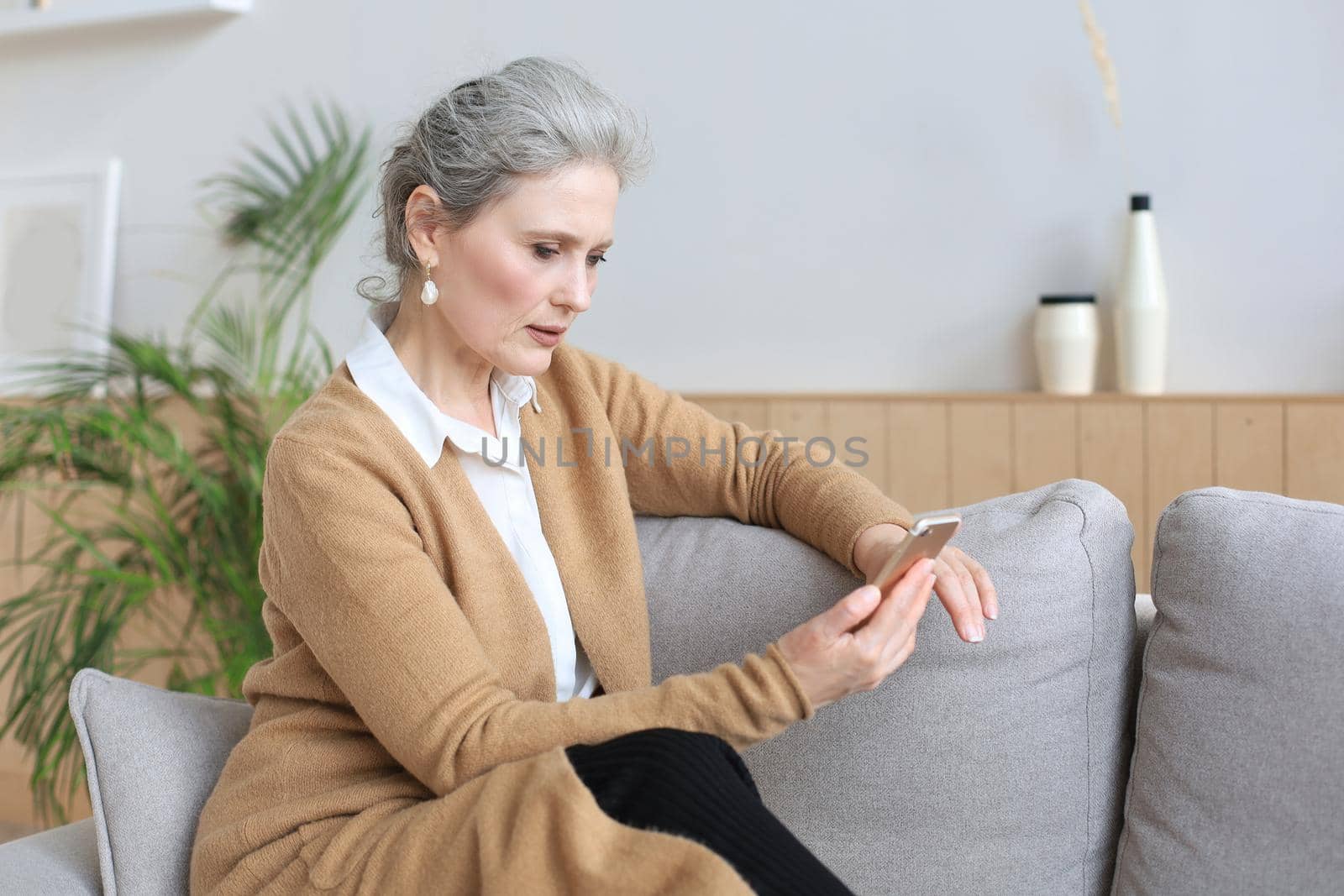 Smiling middle aged woman holding phone, using mobile device apps, looking at screen, while sitting on couch