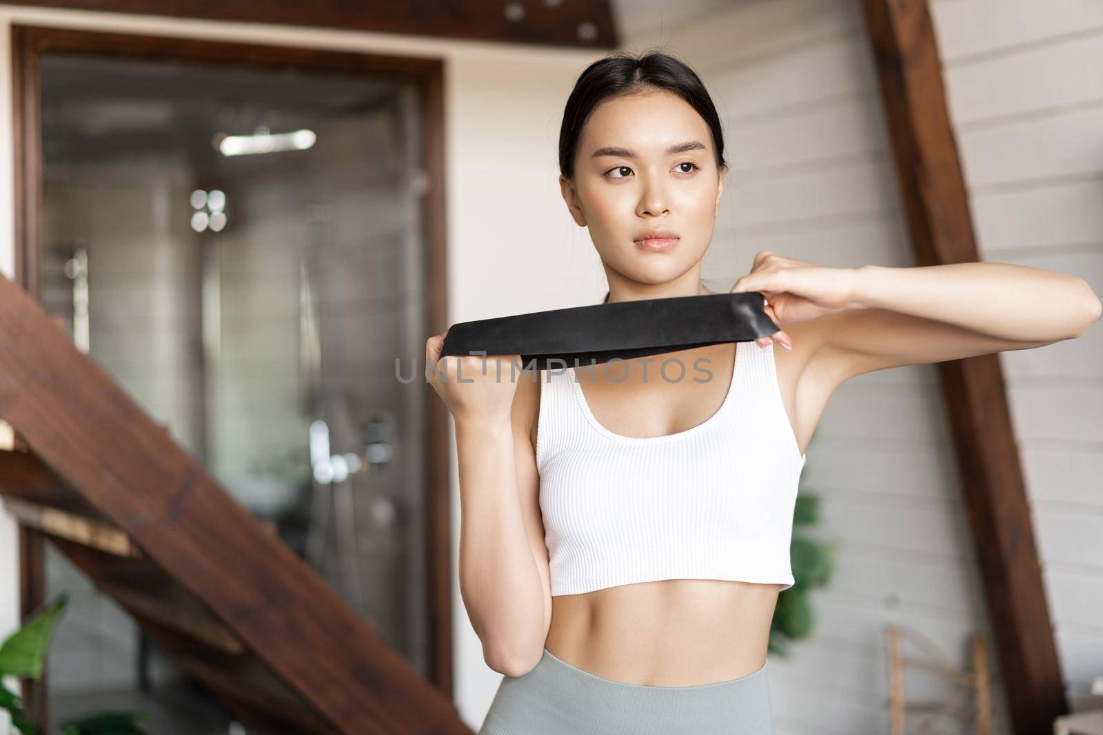 Young asian woman doing fitness workout at home with elastic rope, stretching band for arm biceps, standing in activewear in the living room.