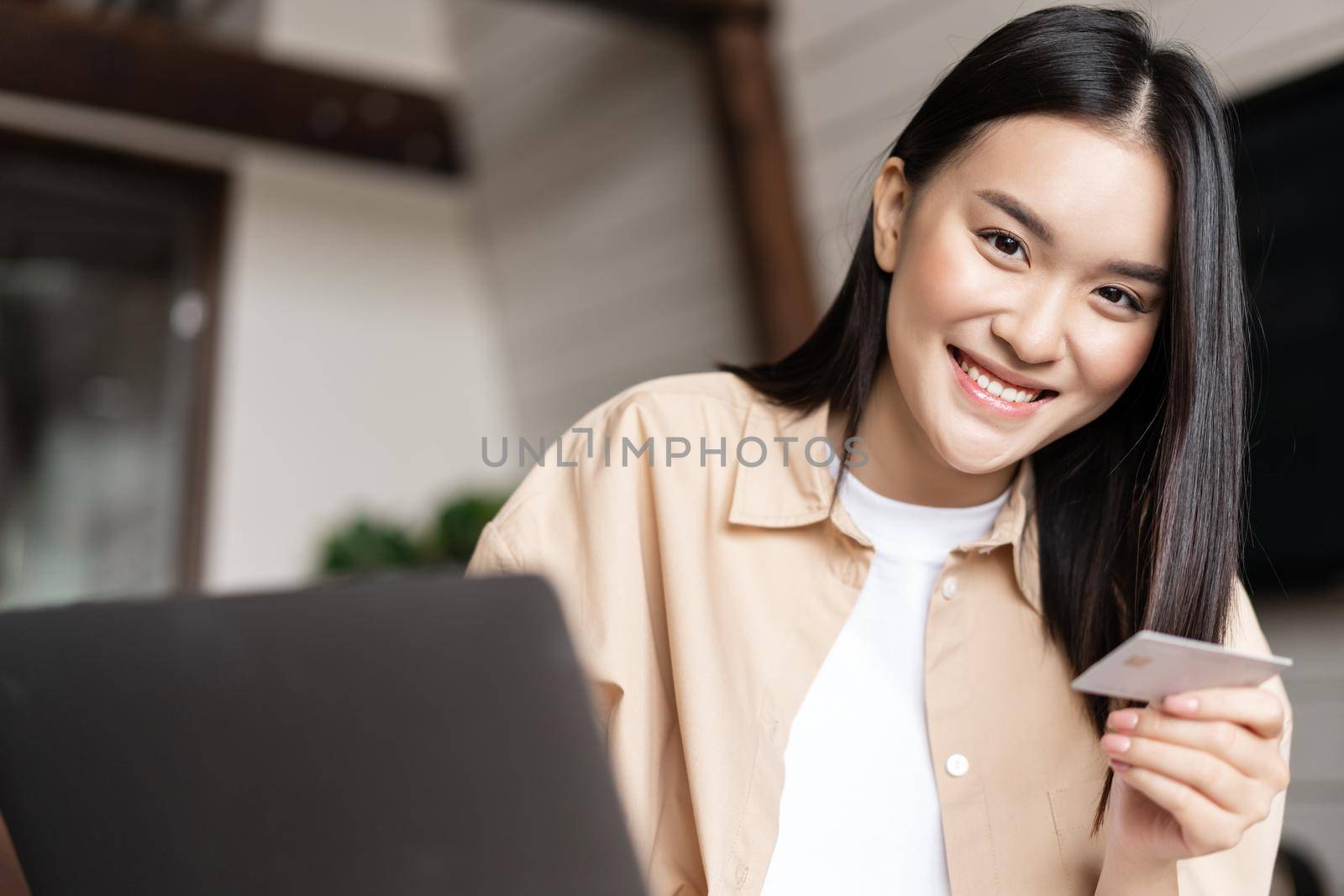 Young asian woman buying online, shopping from her home with use of laptop and credit card, smiling at camera by Benzoix