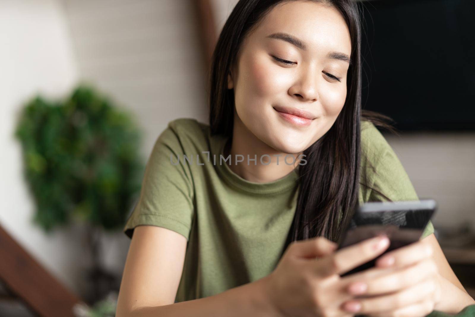 Smiling korean woman looking at mobile phone chat app with dreamy face, sitting at home on couch.