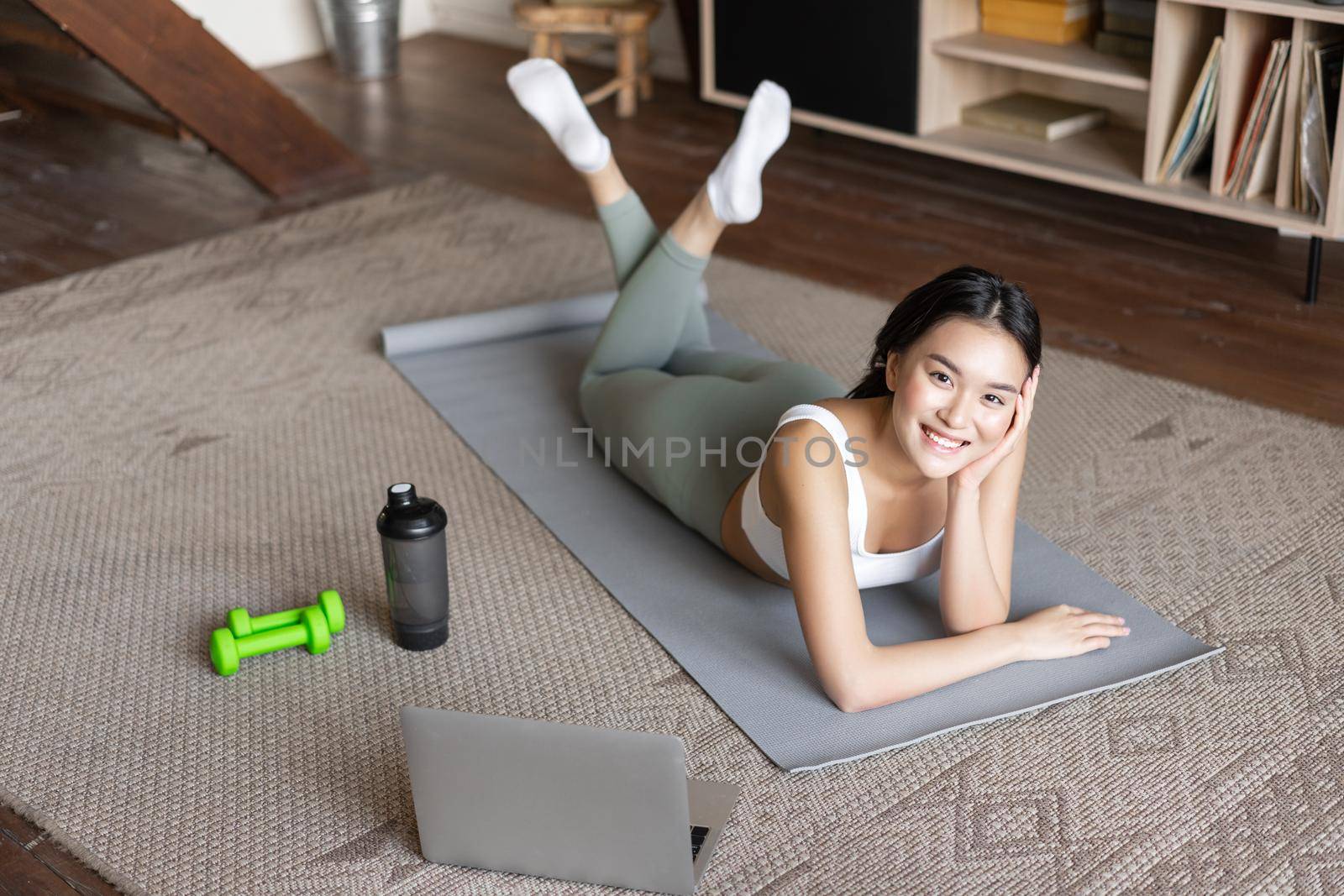 Asian fitness girl having break from workout, exercising at home on floor mat with dumbells and protein shaker bottle, smiling at camera.