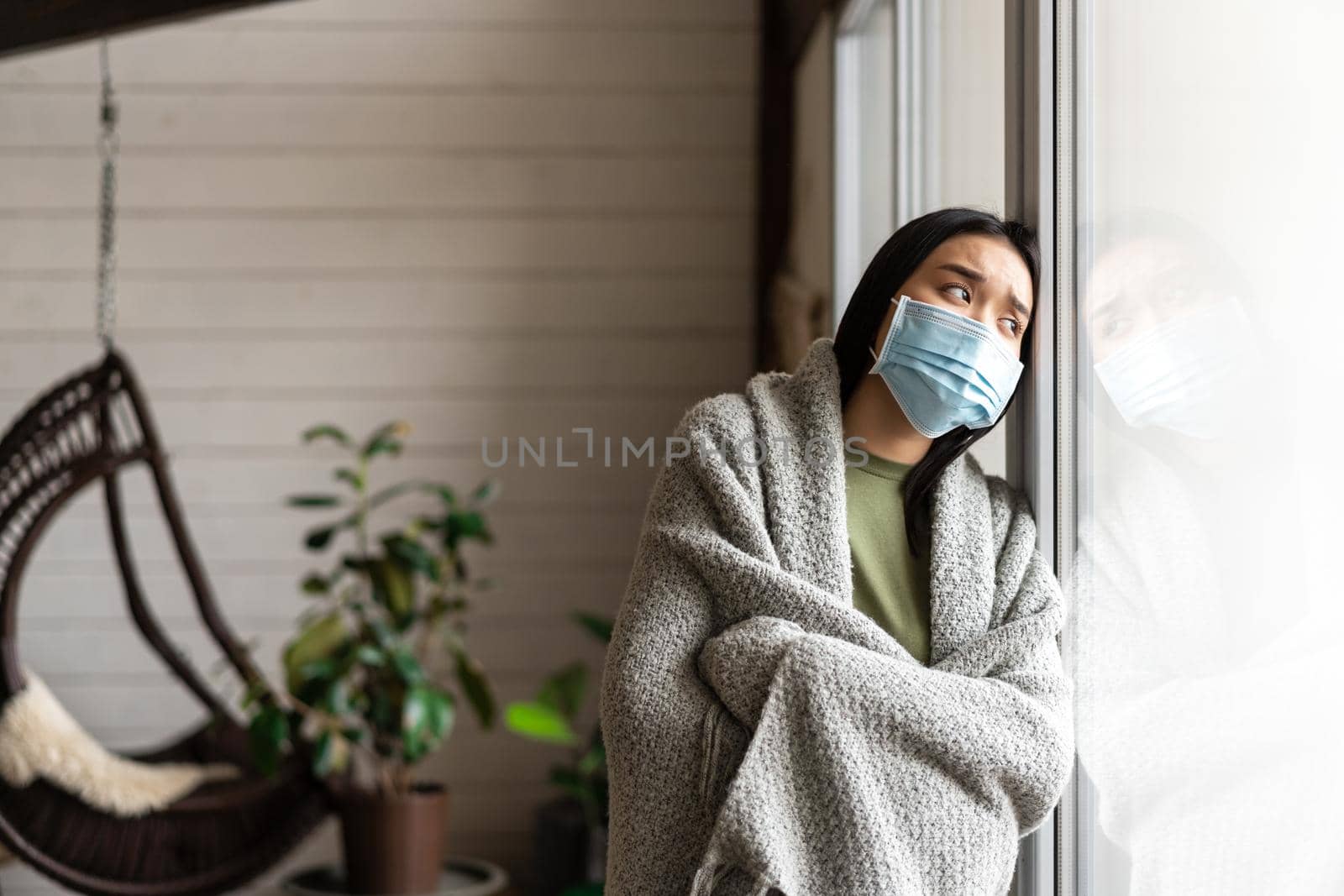 Sick asian woman in medical face mask looking outside window, having flu or covid, staying on quarantine at home by Benzoix