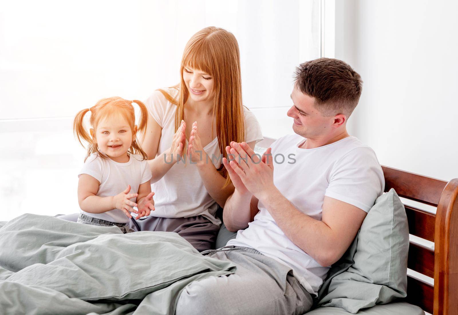 Sunny family morning in the bedroom. Beautiful parents with their daughter staying in the bed and resting. Mother, father and child wearing pajamas smiling