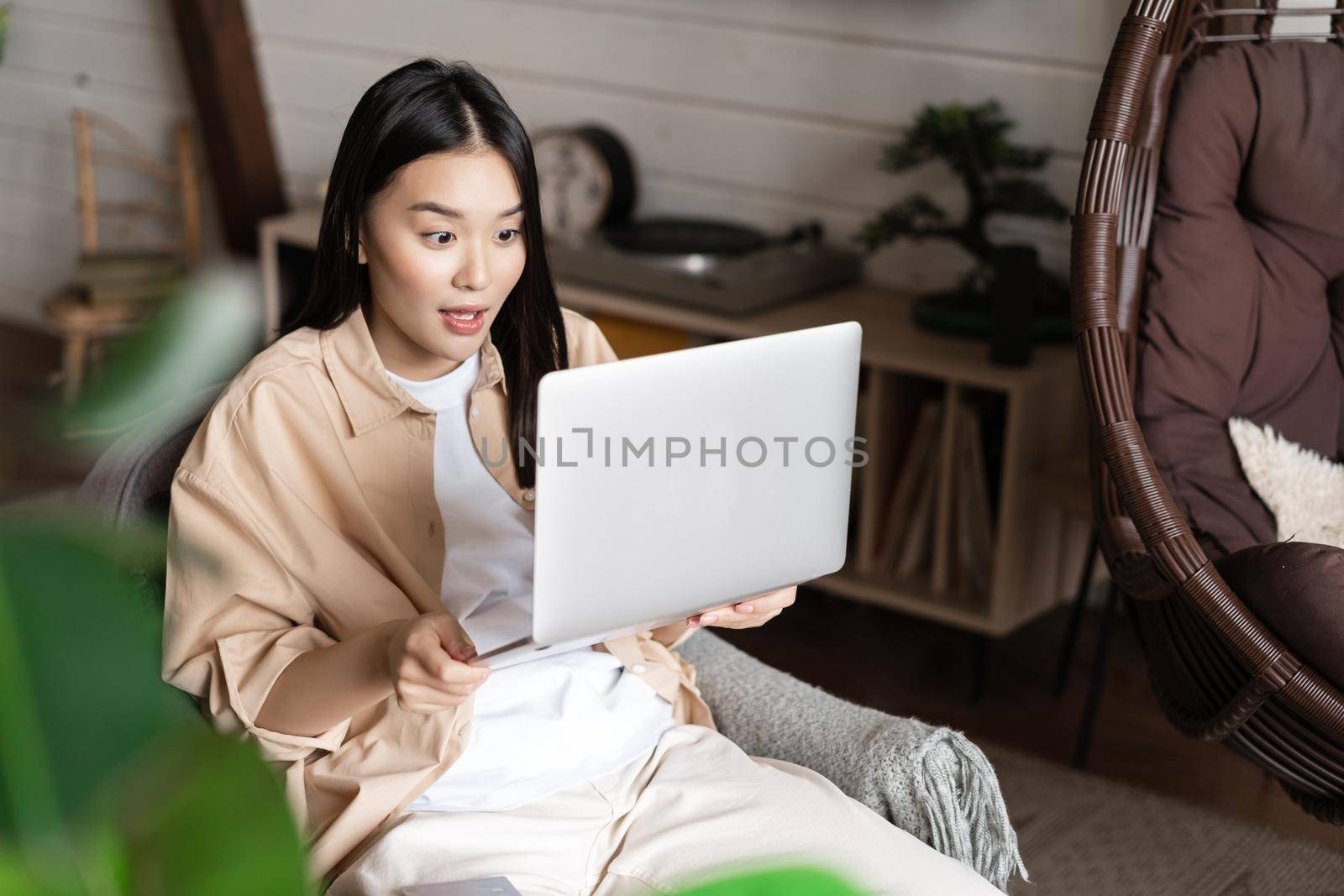 Asian woman looks excited and amazed at laptop screen, winning online, sitting at home by Benzoix