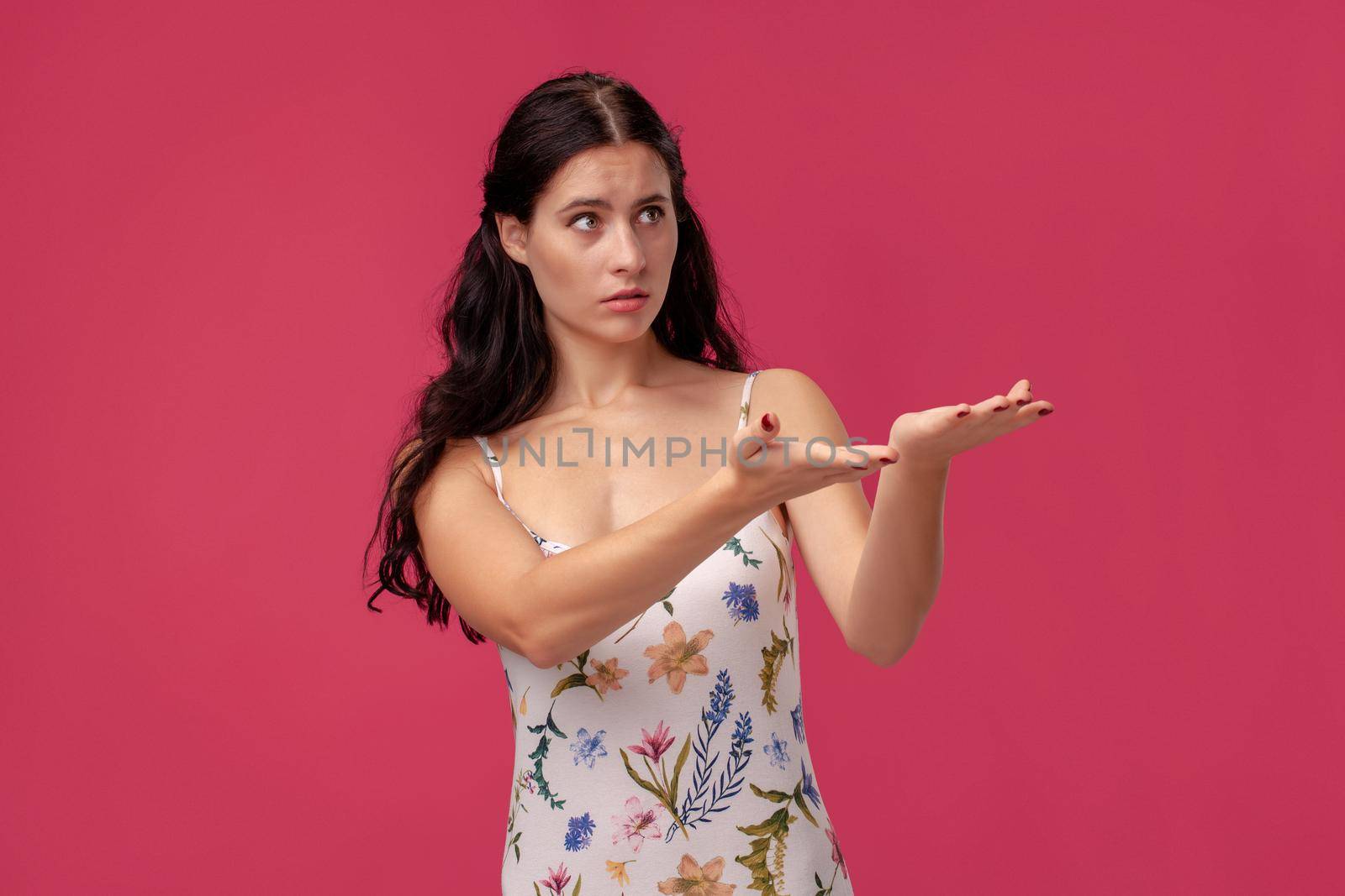 Portrait of a pretty lady in a white dress with floral print standing on a pink wall background in studio. She raised her arms up and looking somewhere. People sincere emotions, lifestyle concept. Mockup copy space.