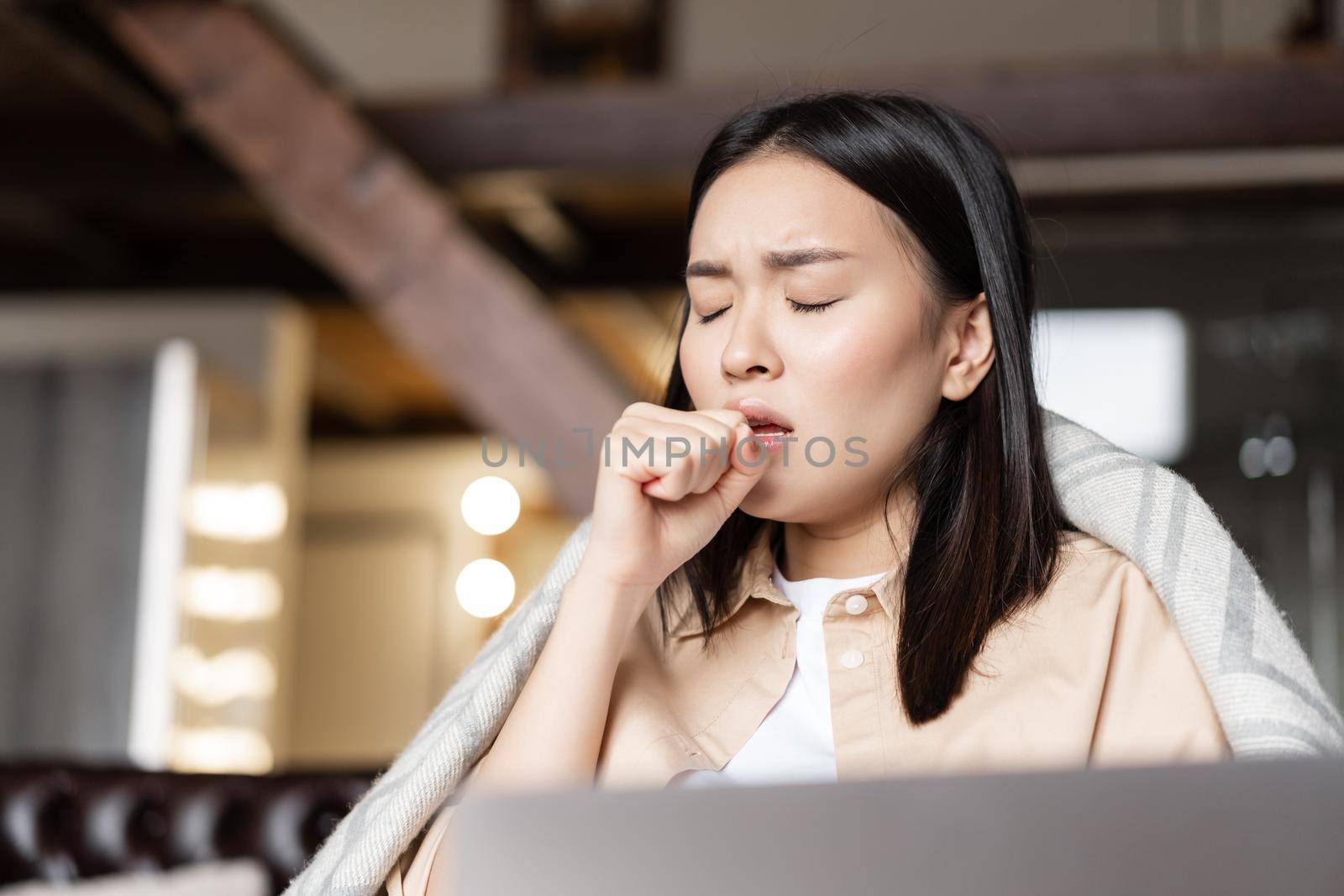 Asian woman being ill, coughing at fist, feeling sick at home, resting with laptop.