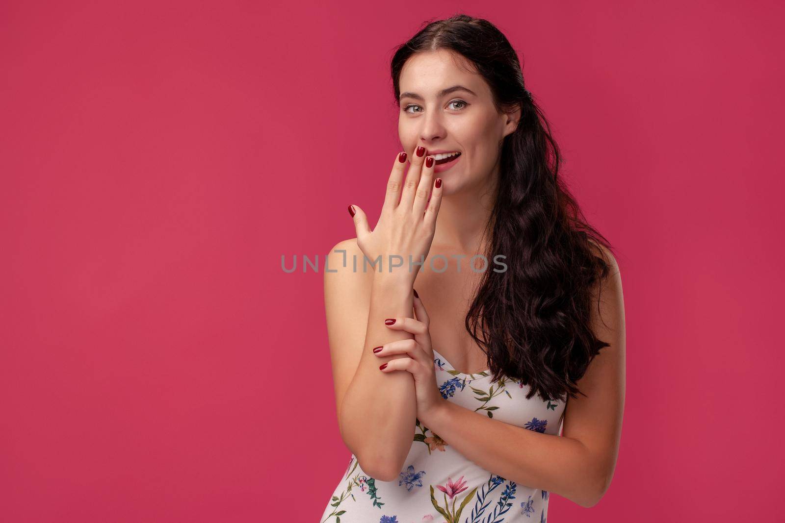 Portrait of a beautiful curly woman, wearing in a white dress with floral print and standing on a pink wall background in studio. She laughs covering her mouth and looking at the camera. People sincere emotions, lifestyle concept. Mockup copy space.