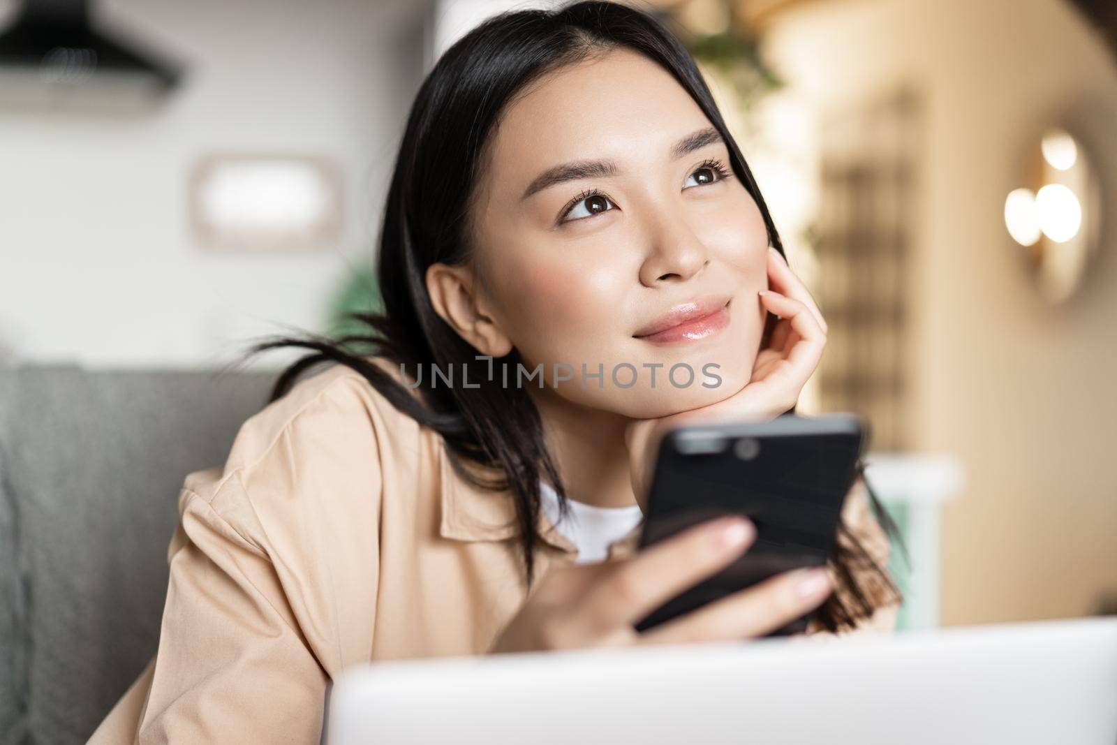Smiling asian girl holding phone and thinking, looking up dreamy, sitting with laptop at home.