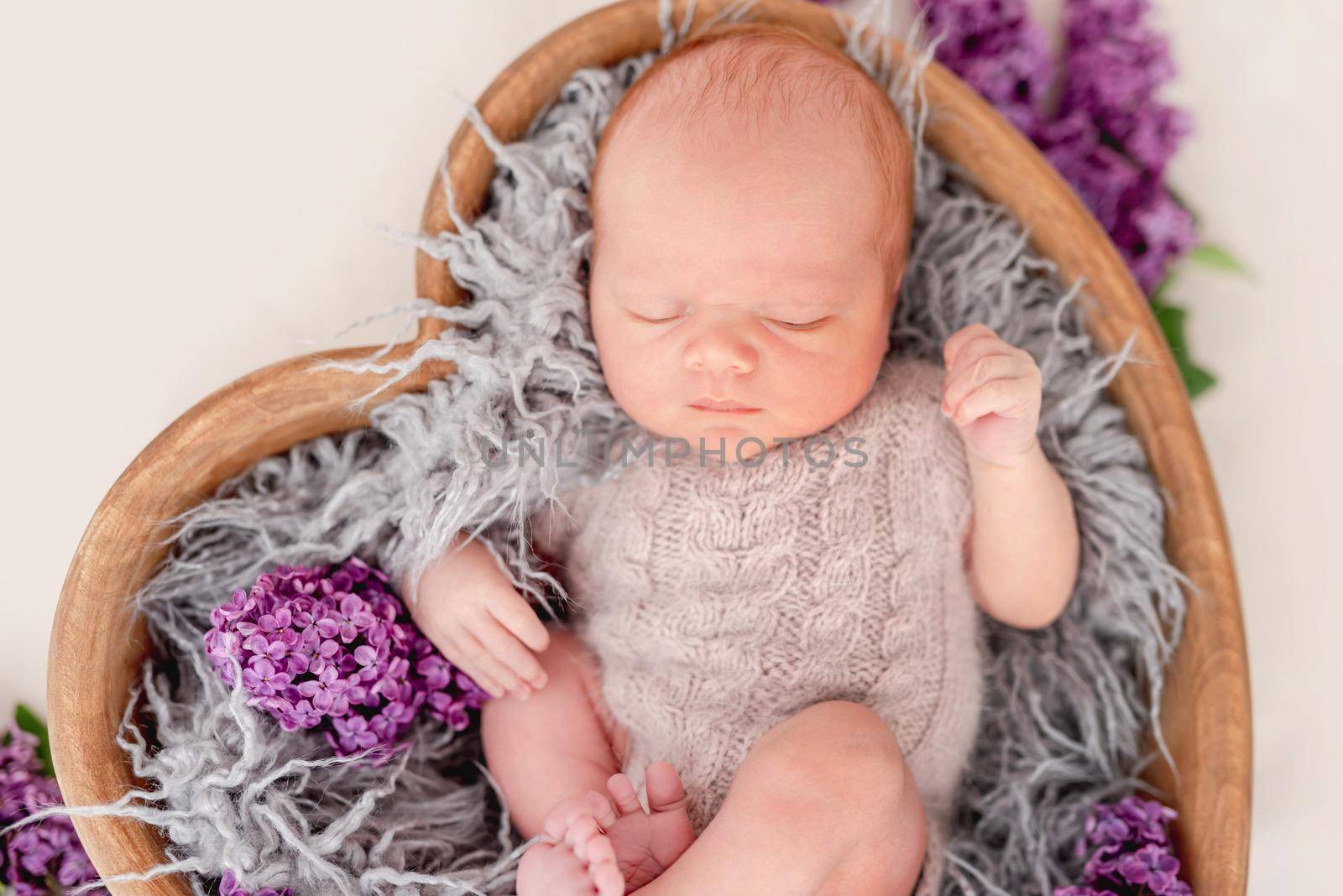 Newborn baby boy wearing knitted beige costume lying in wooden heart shape bed with flowers and sleeping. Adorable infant child napping