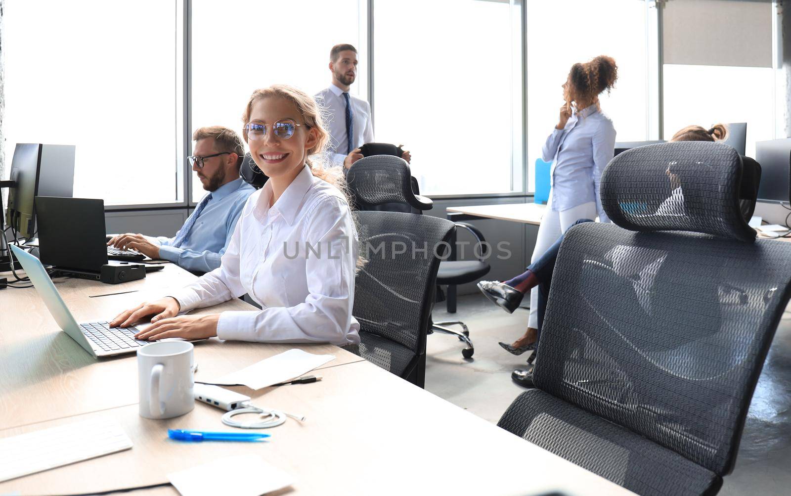 Joyful businesswoman in formalwear working on laptop in the modern office with collegues on the background.
