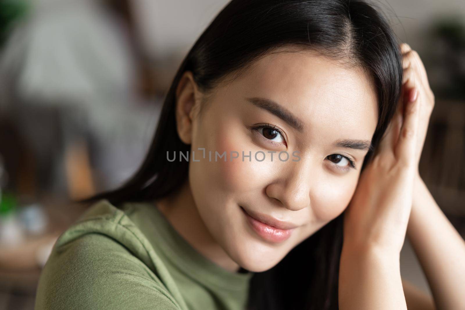 Close up portrait of tender asian girl with glowing natural skin, smiling sensual at camera, sitting at home in cozy clothes.