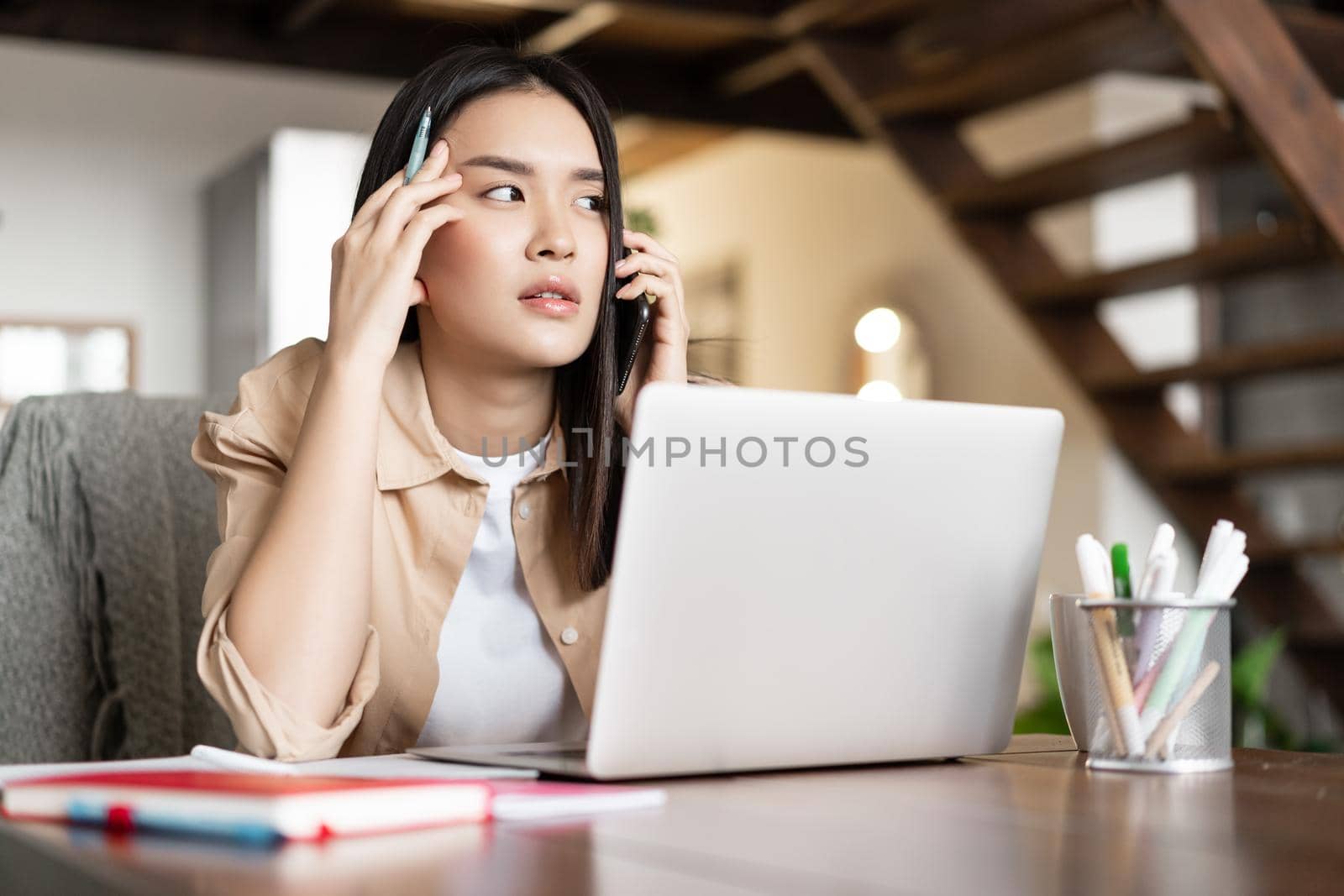 Asian girl answer phone call, looking thoughtful while communicating on phone. Young woman working from home on laptop.