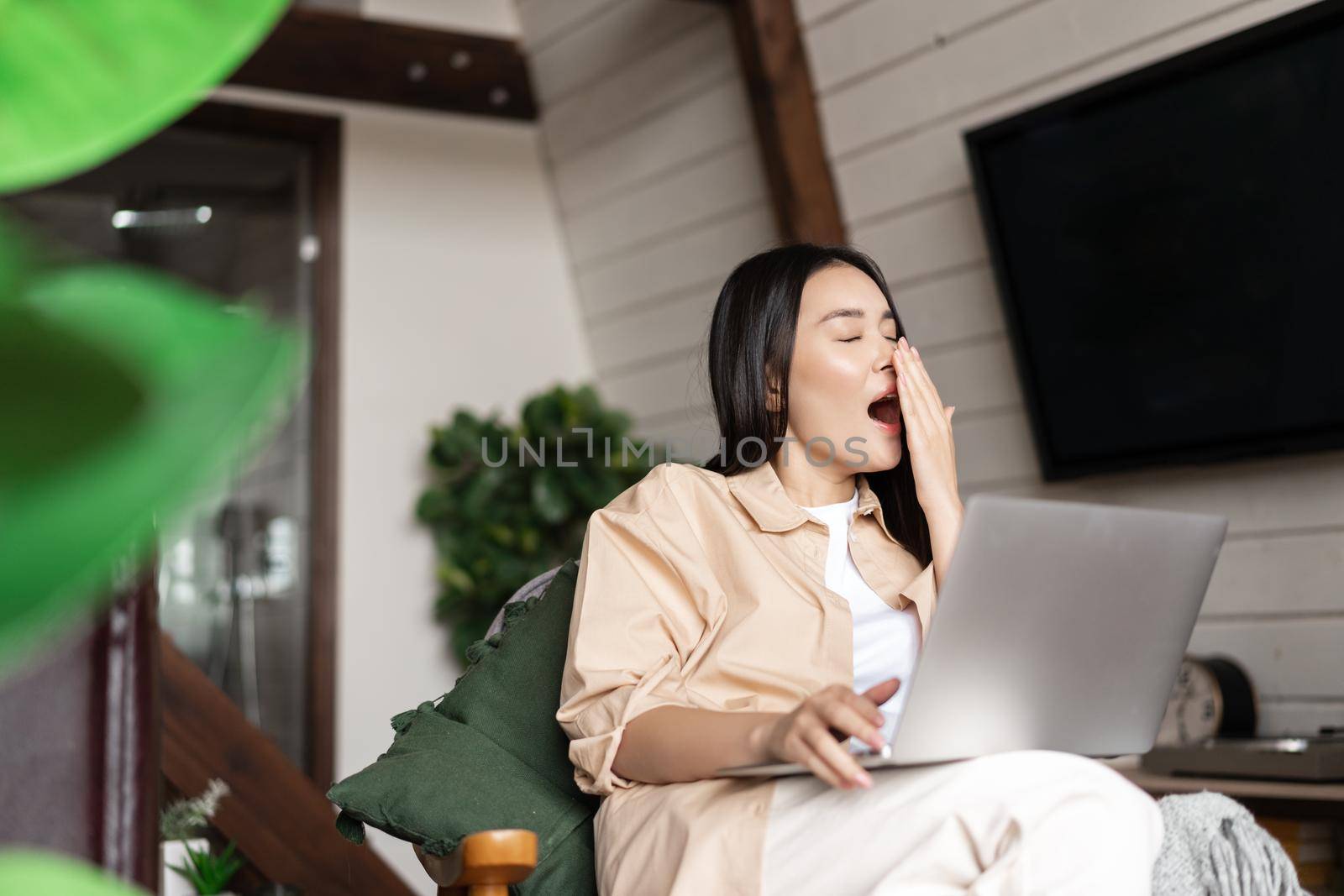 Tired asian girl yawns during work on laptop, sits at home, works remotely from her living room by Benzoix