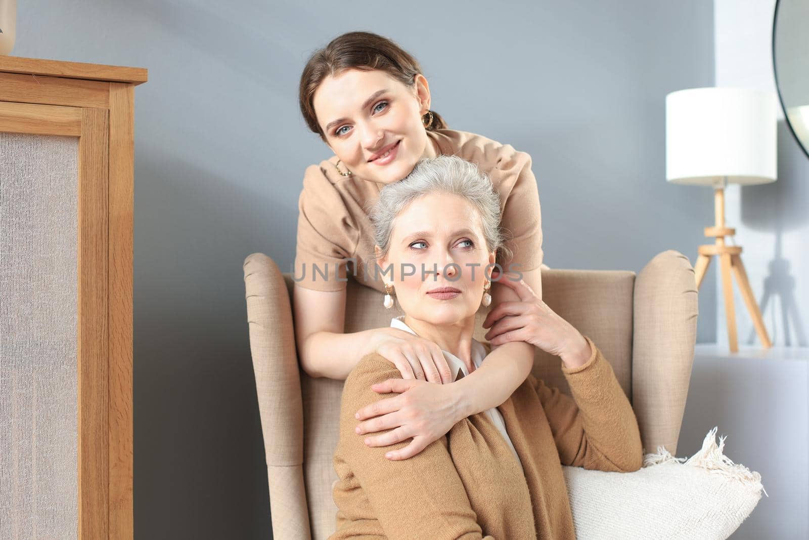 Happy daughter hugging older mother, standing behind chair in living room, enjoying tender moment at home