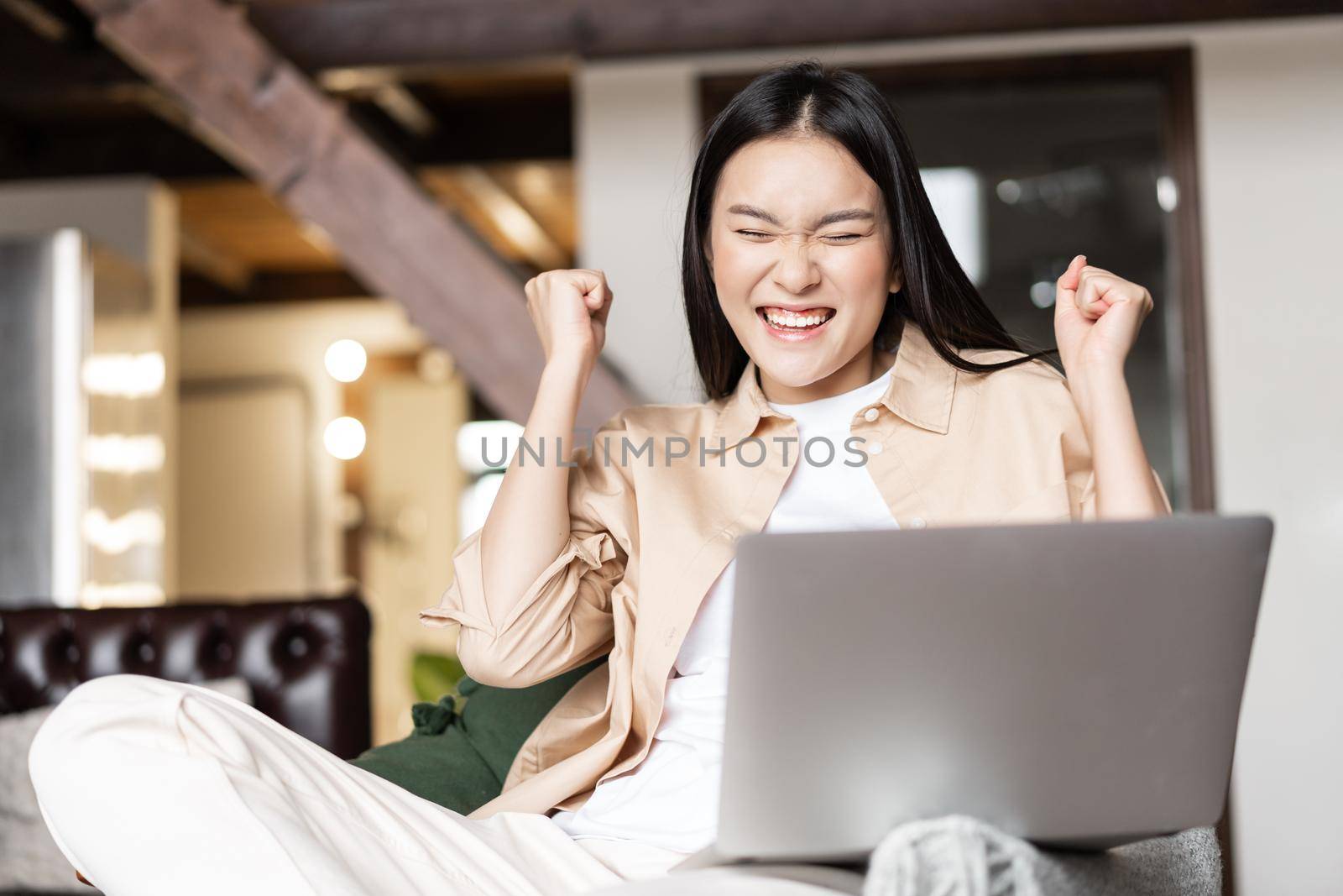 Asian woman celebrating, cheering from good news on laptop, triumphing from joy, sitting in living room at home.