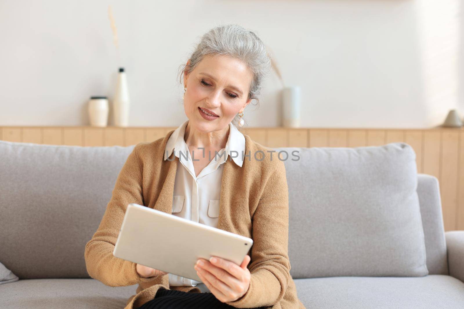 Cheerful middle aged woman sitting on sofa, using computer tablet apps, looking at screen,reading good news in social network, shopping or chatting online