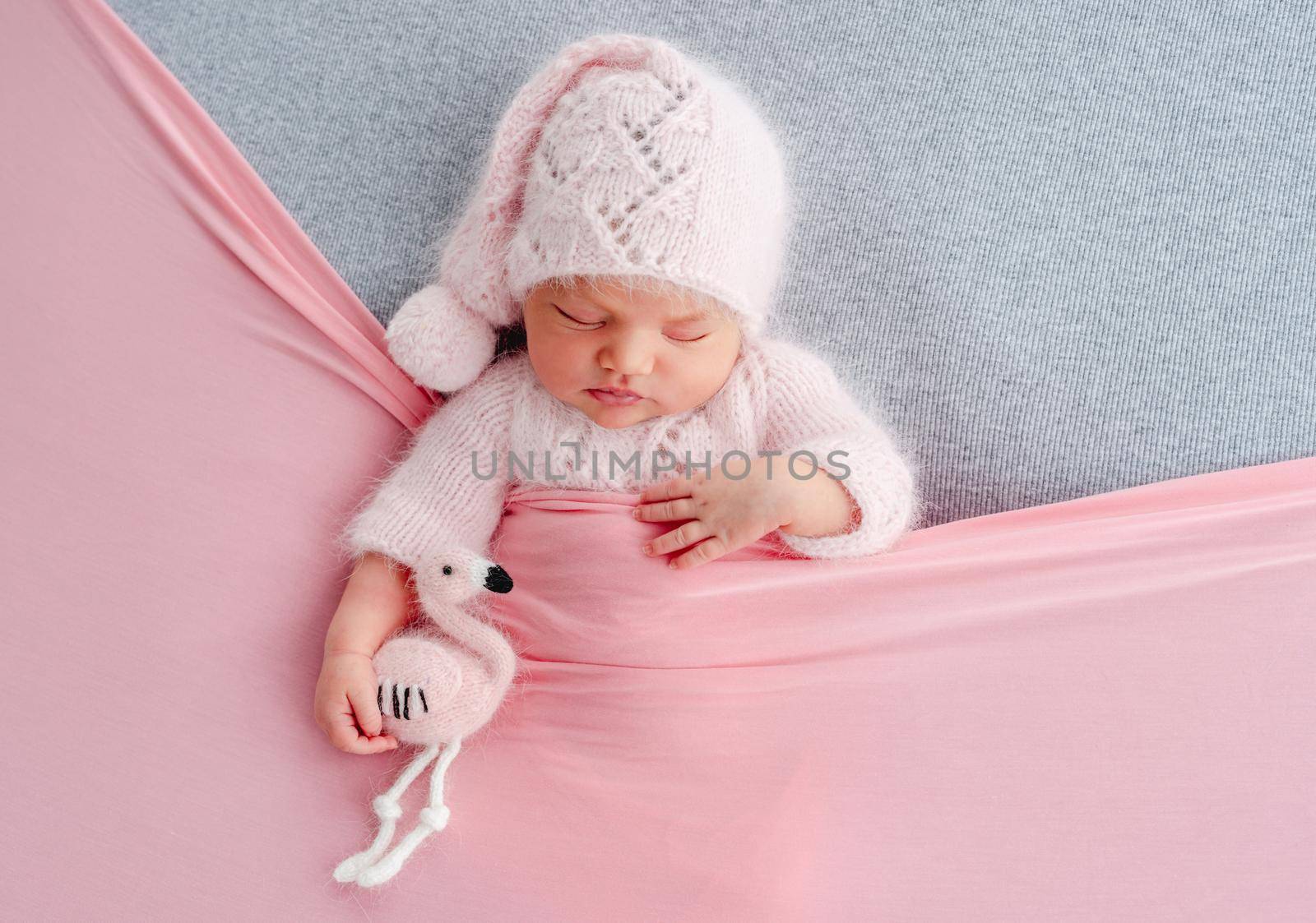 Cute newborn baby girl in knitted hat sleeping on grey stylized background and holding flamingo toy in her hands. Adorable infant child napping under pink blanket during studio photoshoot