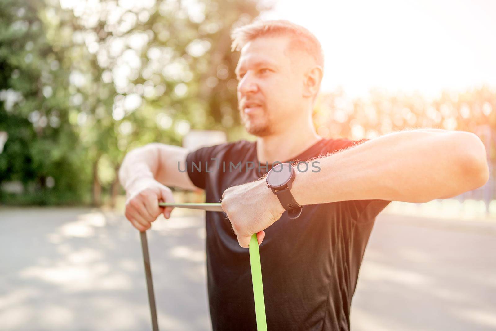 Sportive man guy training with elastic rubber band at the stadium outdoors with sunlight. Athlete male person during workout with additional sport equipmant