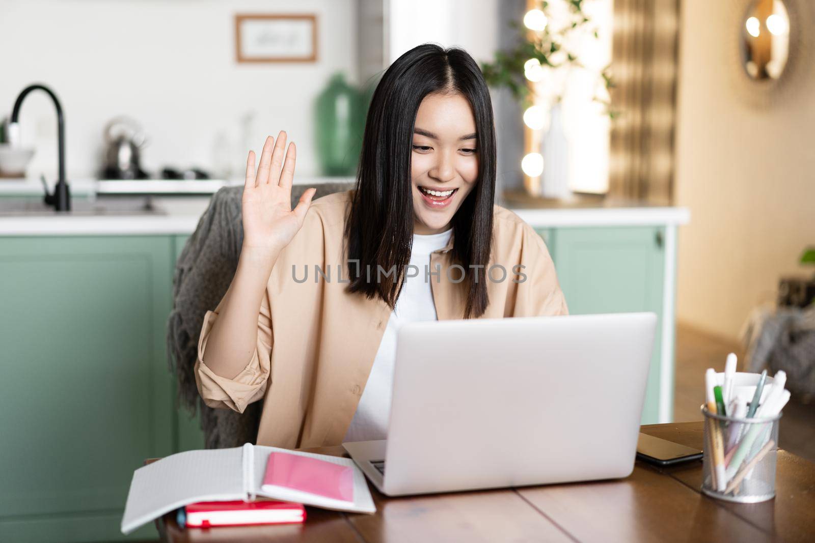 Smiling asian woman working from home, having call online video meeting with coworkers, waving hand at laptop and saying hello. Student video chatting, studying remotely.