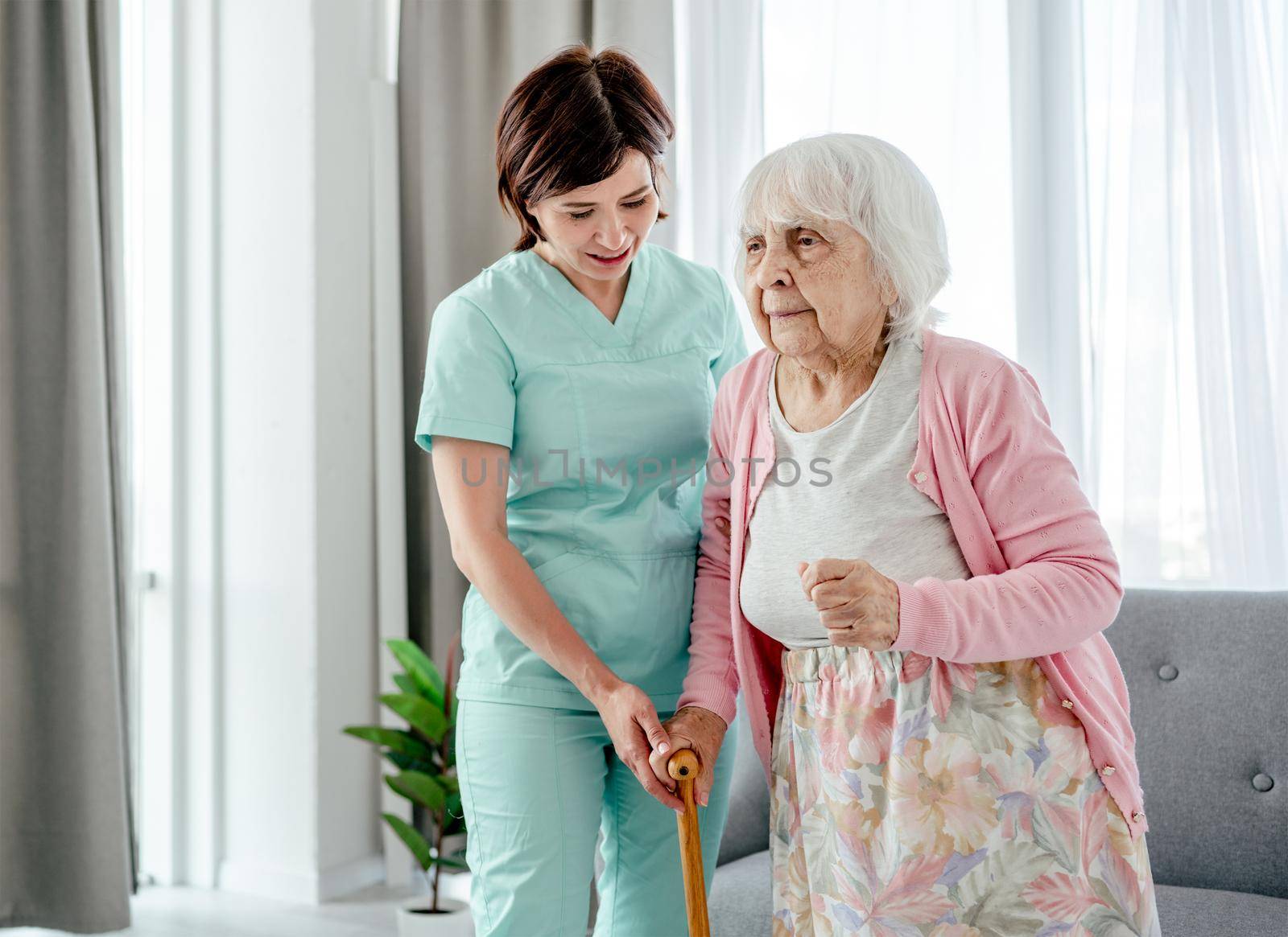 Young nurse helps elderly woman at home. Healthcare worker girl cares about senior female person indoors