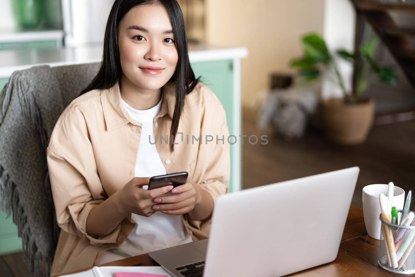 Smiling asian woman working with laptop from home. Girl student holding smartphone, sitting near computer and looking at camera.