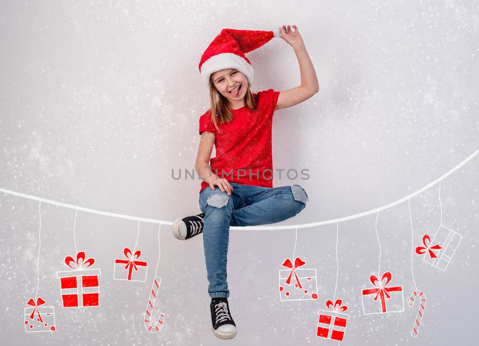 Little girl in santa hat sitting on chalk drawn thread with hanging christmas presents on white wall background