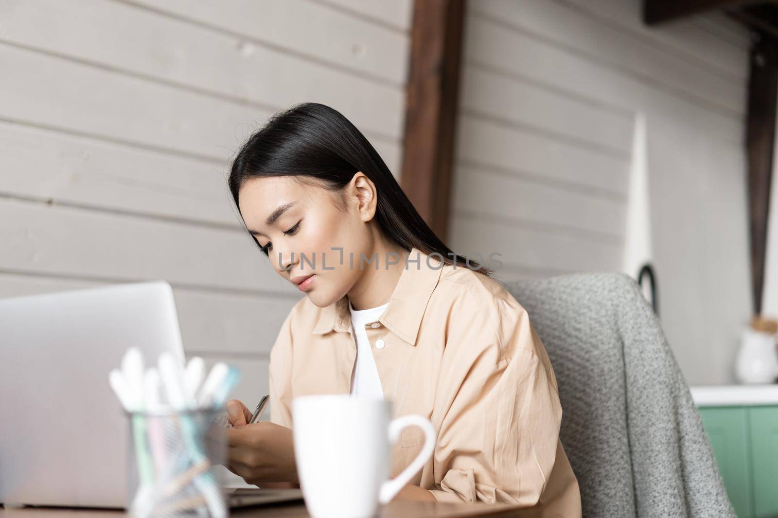 Young woman working on laptop at home. Girl writing down homework and listening webinar on computer, attend online lecture.