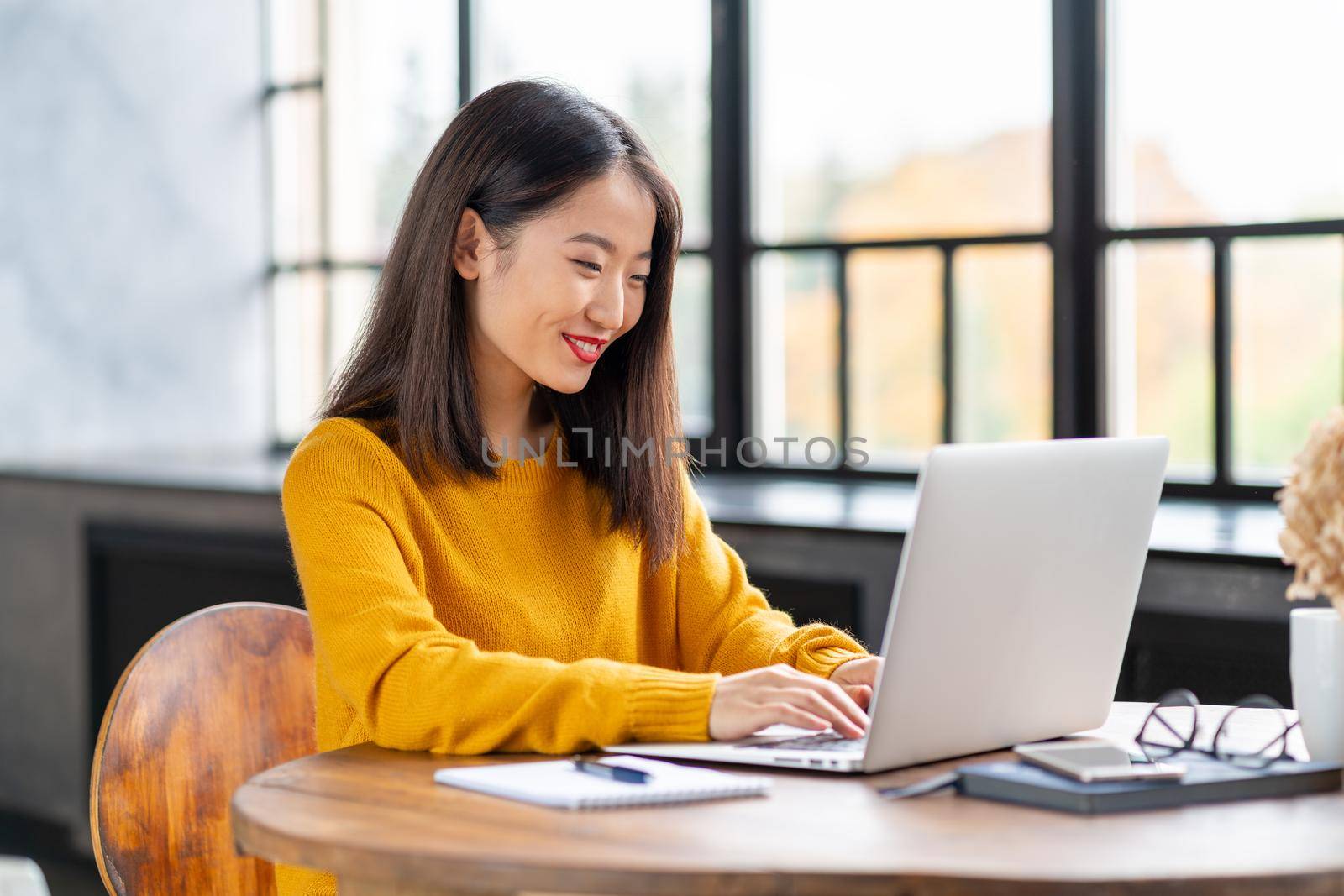 Asian woman working on laptop at home or in cafe. Young lady in bright yellow jumper sitting at desk typing on computer. Business oriental female in front of window