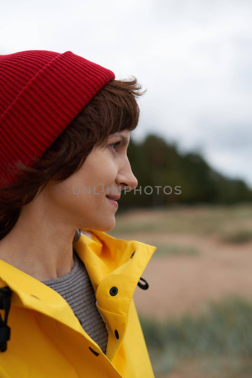 One with nature. Crop closeup portrait of mature beautiful woman in bright yellow jacket and red hat on shore of North sea on autumn day. Mood Scandinavian weather.