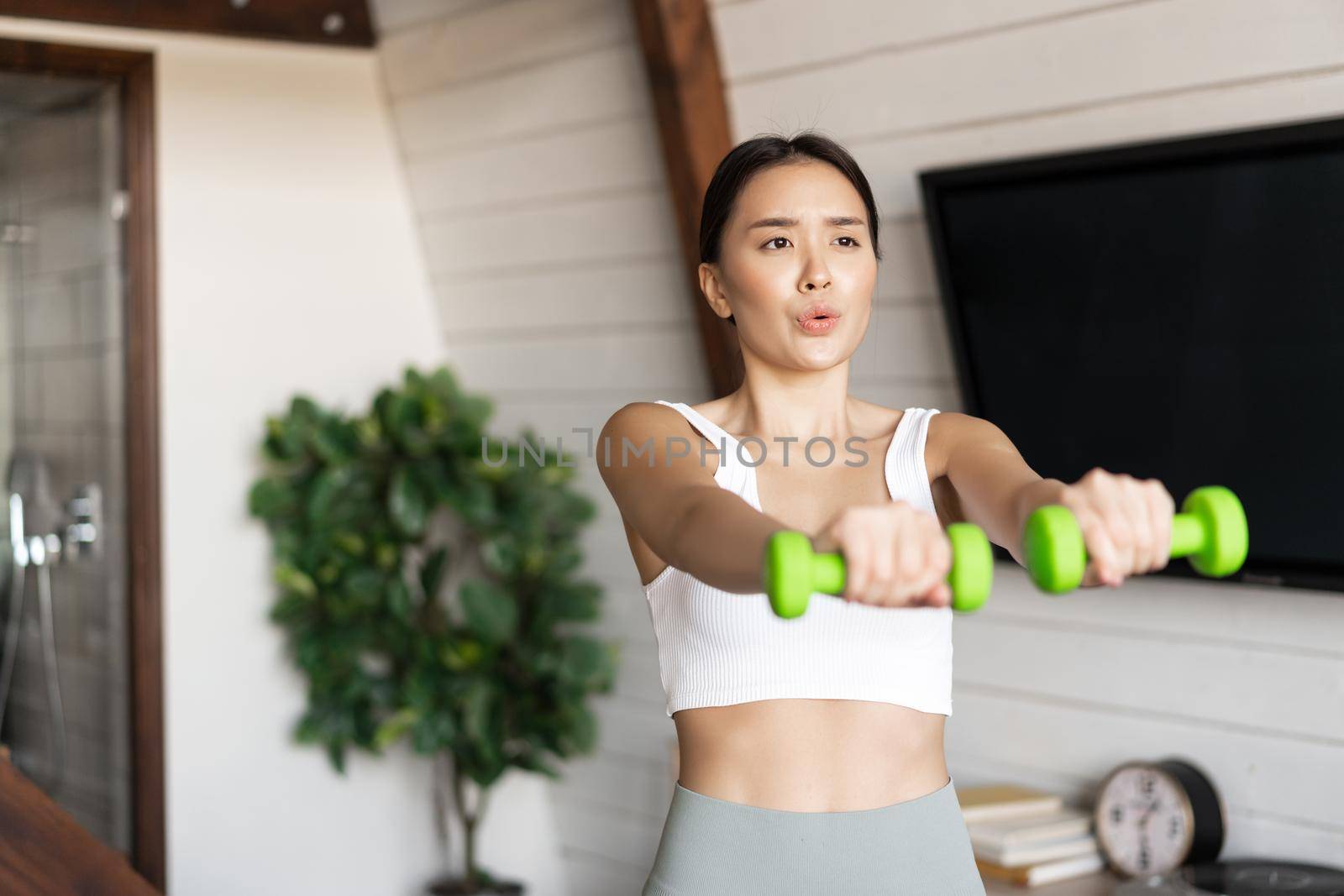 Fitness asian girl doing workout at home in living room, lifting dumbbells, wearing activewear for sport activities.