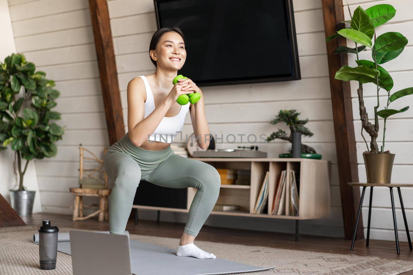 Young asian woman doing workout at home with dumbbels, making squats on floor mat, follow online fitness instructor guide on laptop.