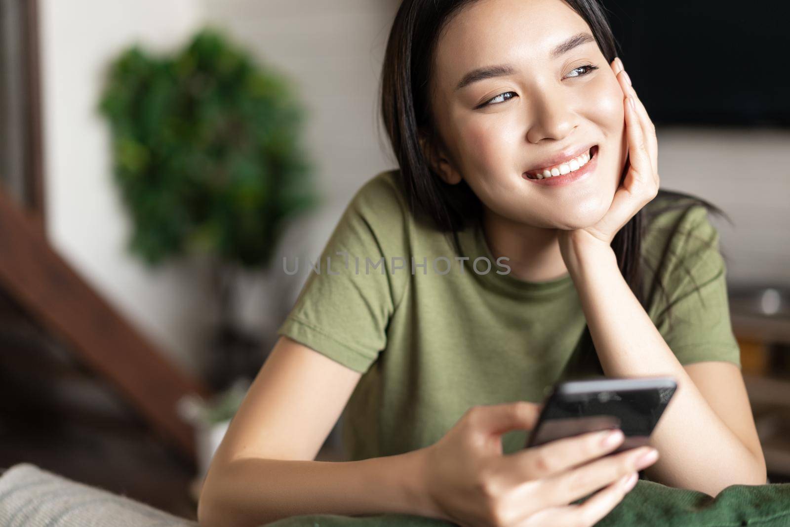 Young asian woman placing an order, waiting for delivery on mobile phone app, sitting at home and looking outside window.
