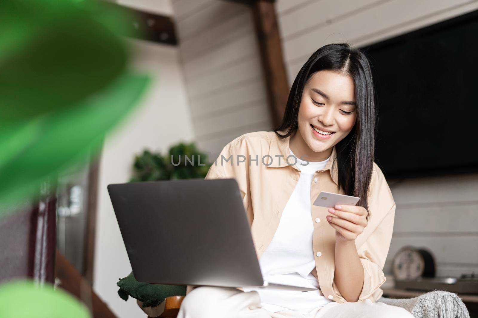 Smiling asian girl paying for online shopping on laptop, looking at her credit card, sitting at home in living room with computer by Benzoix