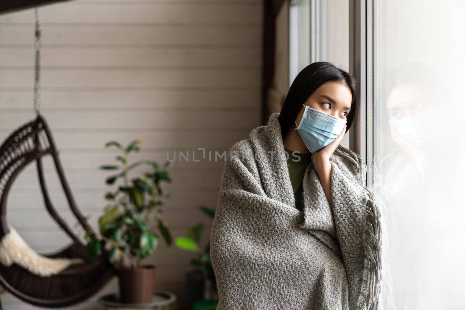 Sad sick asian girl on quarantine, standing near window at home and looking outside, wearing medical face mask from covid.