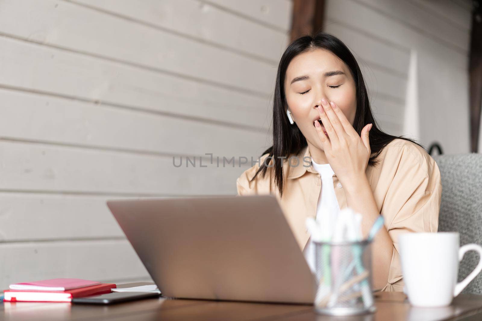 Tired girl student working on laptop and yawning, drinking coffee while studying, doing homework on computer.