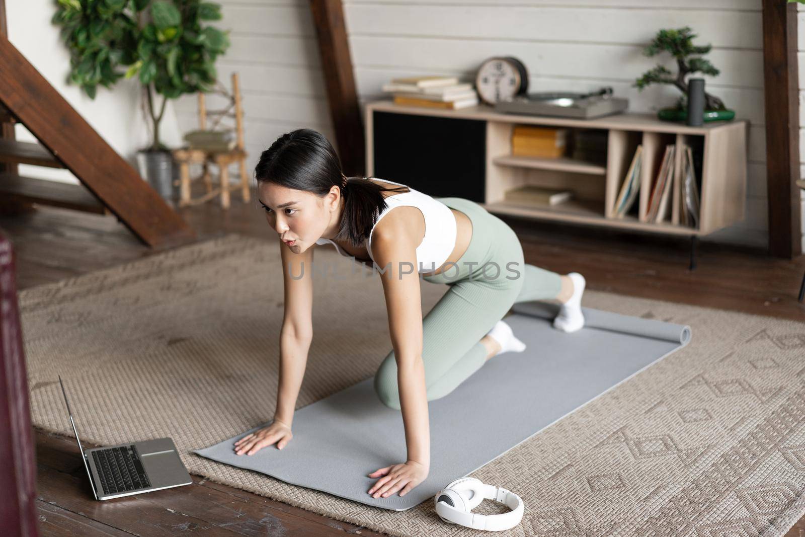 Image of young asian girl doing workout at home, following fitness instructor online on laptop, doing exercises in living room, wearing activewear.