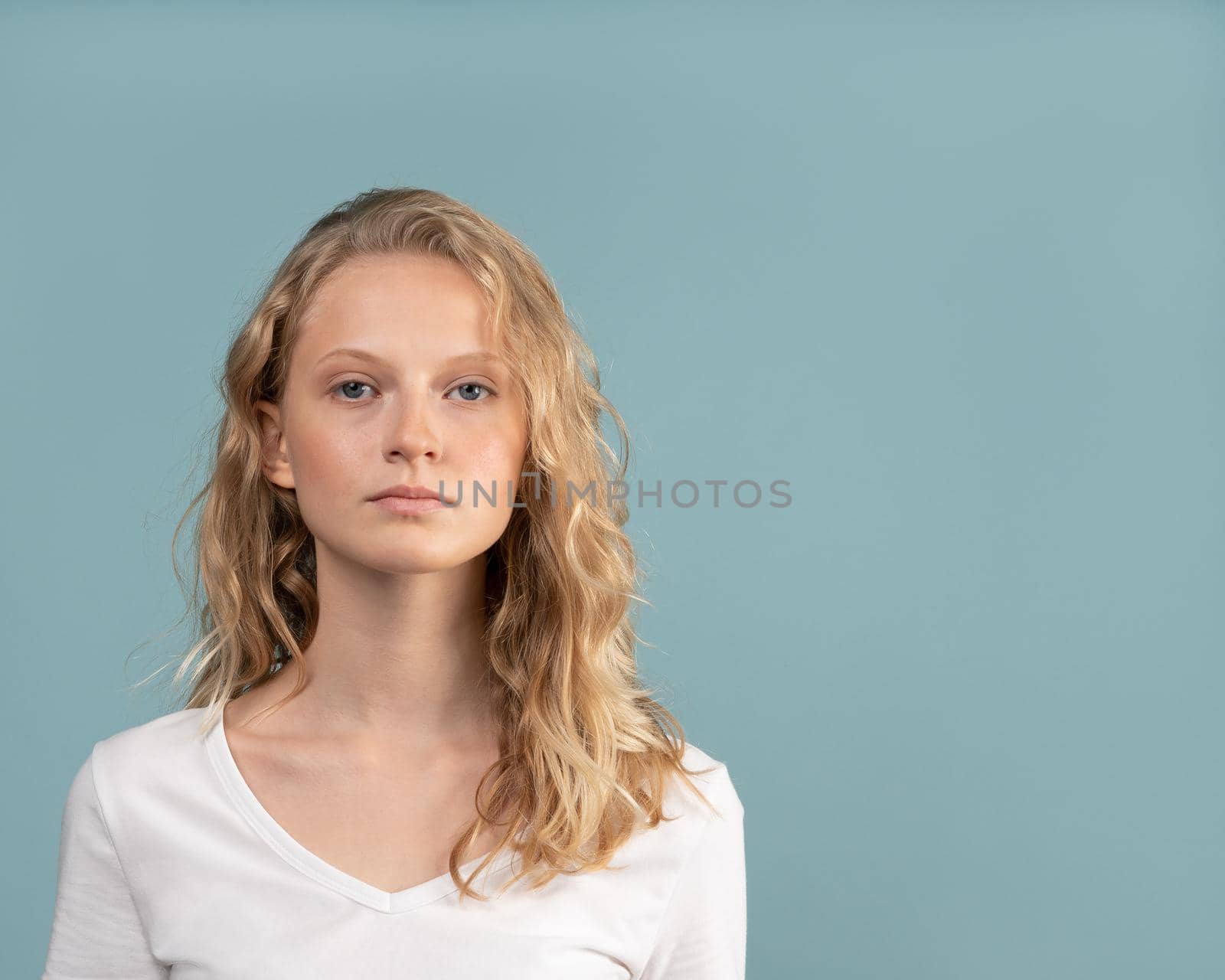 Beautiful young serious clever blonde woman without makeup on color neutral tone Aqua Menthe wall. Pretty female with curly hair in white t shirt