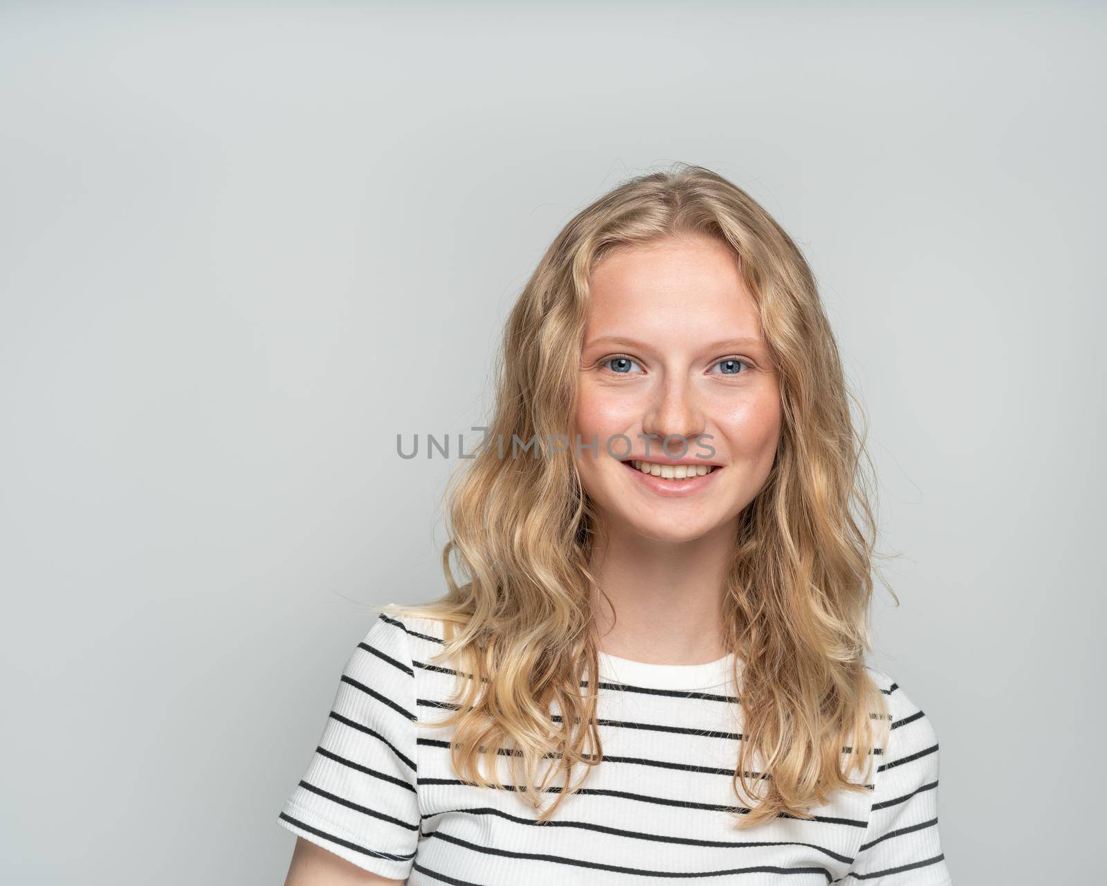 Beautiful young smiling clever blonde woman without makeup on white wall. Pretty female with curly hair in white t shirt