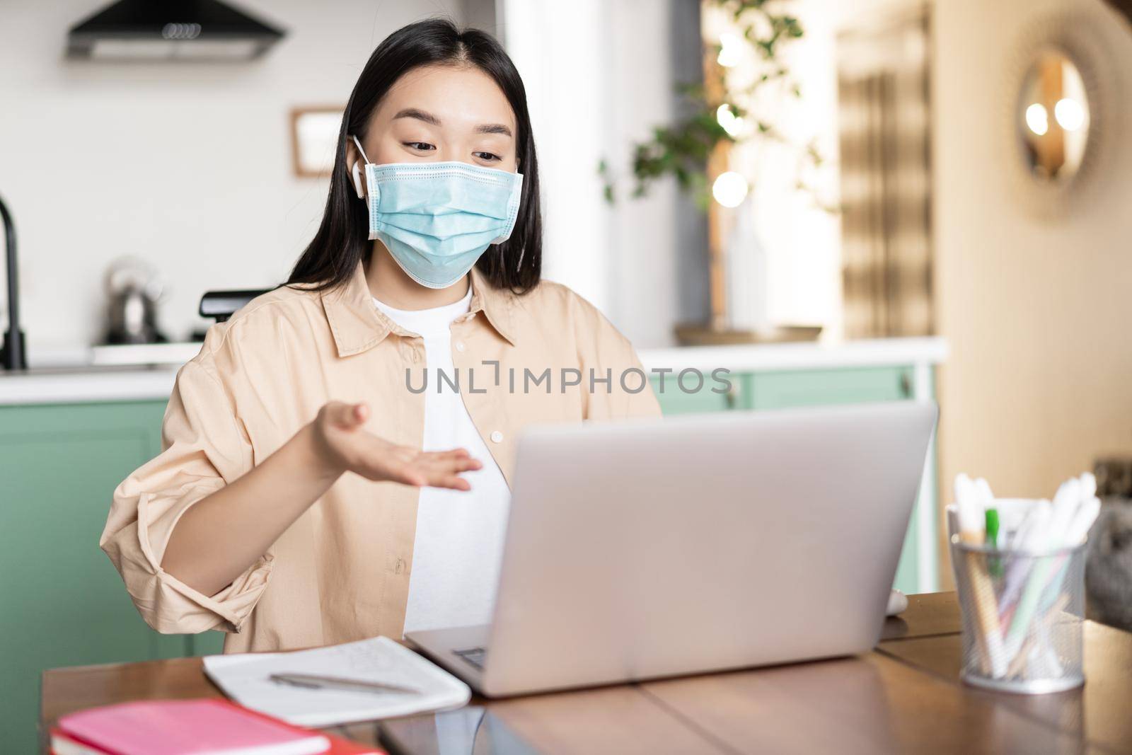 Young asian girl talking at laptop in medical mask, sitting at home, work or study remotely by Benzoix