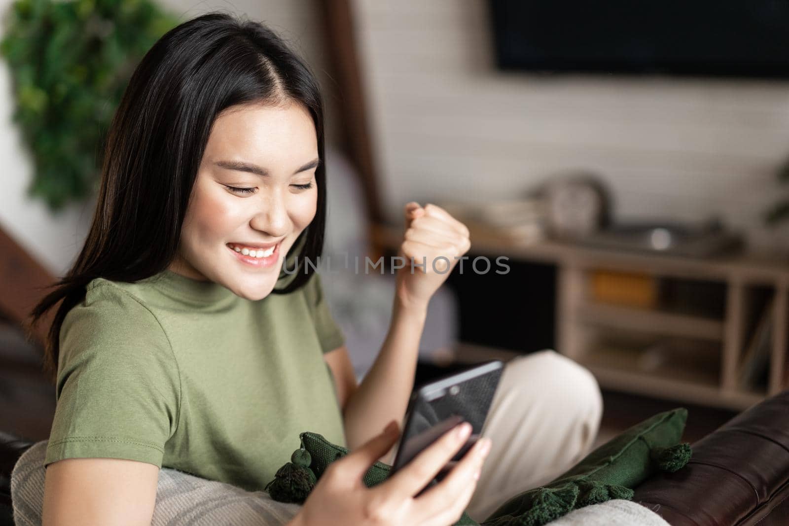 Excited smiling asian girl winning on mobile phone, saying yes and rejoicing, looking at smartphone screen pleased.